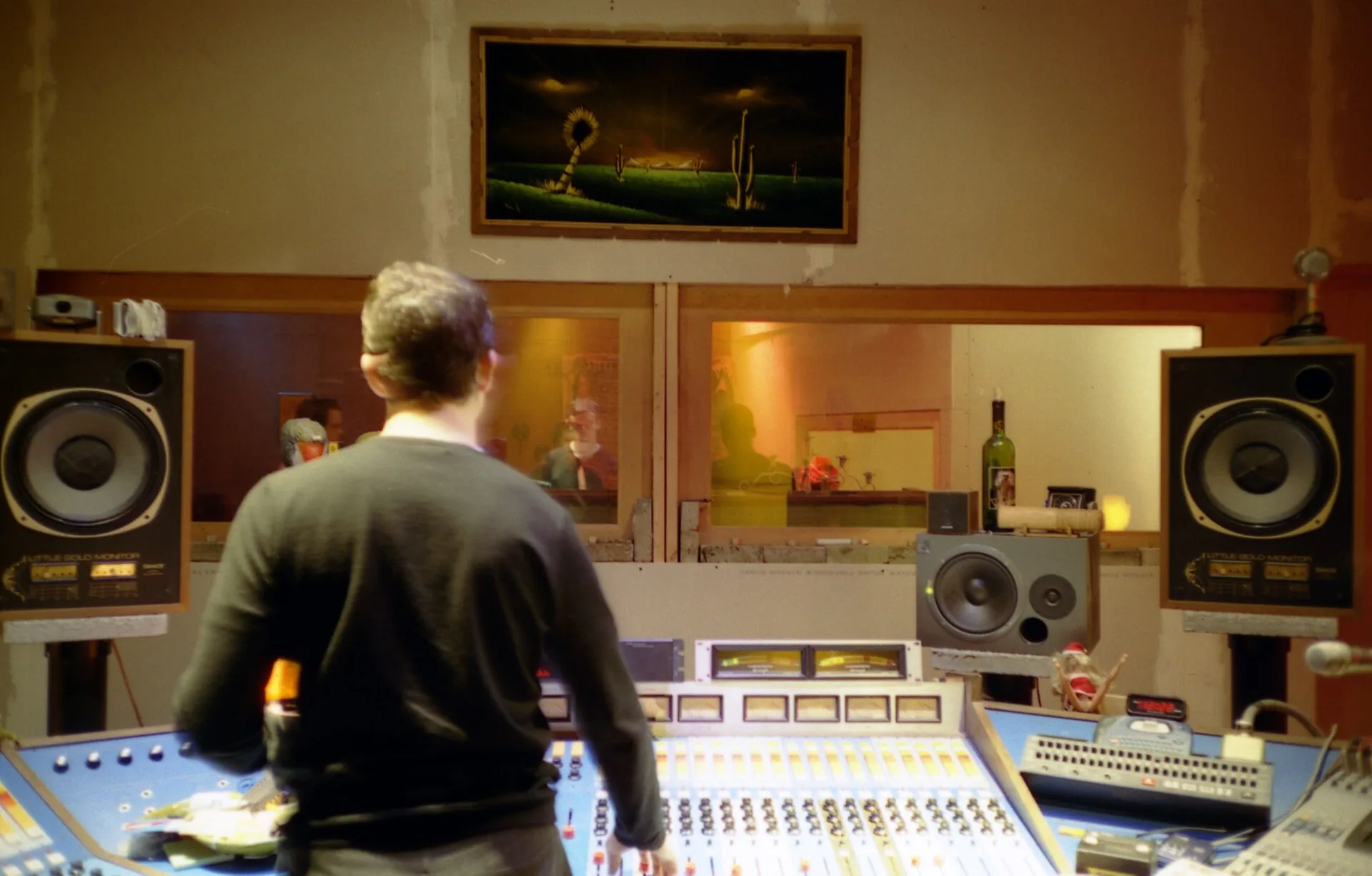 An audio engineer in the control room of a professional sound recording studio. In front of him is the mixing console with a plurality of linear potentiometers for regulating the recording level of the different instruments/voices playing in the live room behind the glass window. In front of the eng. Music Studio Compliance Checklist: Opening Stages